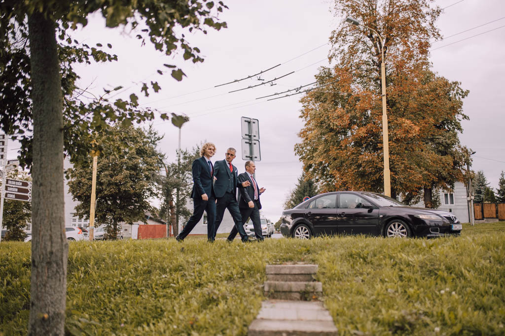 Také pekné - svadobný fotograf - Košice, Prešov Traja muži v oblekoch kráčajú po chodníku vedľa trávnatej plochy, v pozadí je čierne auto a stromy. Je zamračený deň a zdá sa, že muži sú zaujatí rozhovorom.