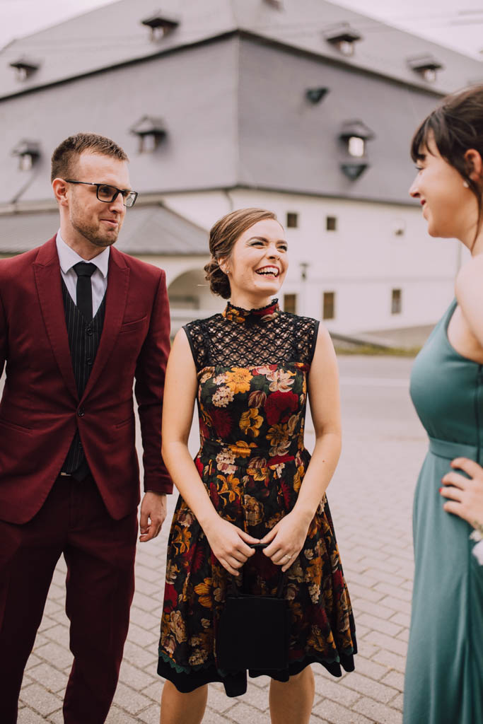 Také pekné - svadobný fotograf - Košice, Prešov Vonku stoja traja ľudia vo formálnom oblečení, usmievajú sa a rozprávajú. Pred veľkou budovou s vikiermi stoja muž vo vínovom obleku a dve ženy vo vzorovaných šatách a v šatách v talianskej farbe.