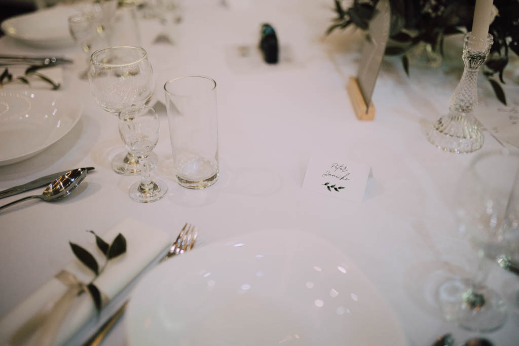 Také pekné - svadobný fotograf - Košice, Prešov Detailný záber na elegantné prestretý stôl s bielymi taniermi, pohármi, strieborným príborom, zrolovaným obrúskom so zeleňou a malou kartičkou s nápisom Päťdesiat hostí na bielom obruse.