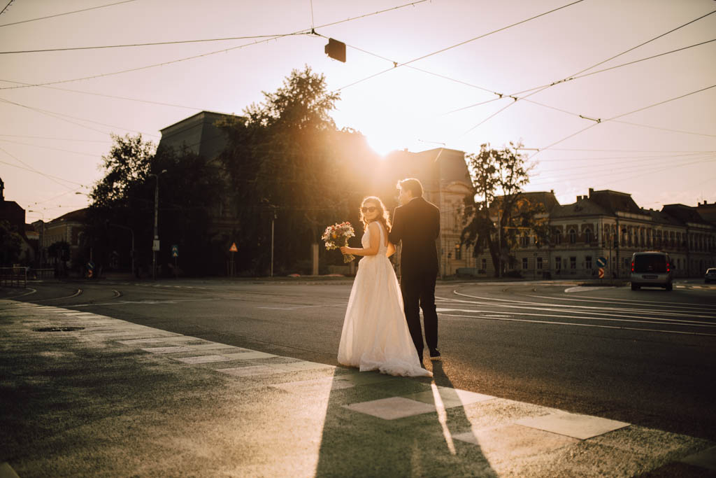 Také pekné - svadobný fotograf - Košice, Prešov Nevesta a ženích kráčajú ruka v ruke po mestskej ulici pri západe slnka, nevesta drží kyticu a slnečné svetlo prúdiace za nimi vrhá dlhé tiene na chodník.