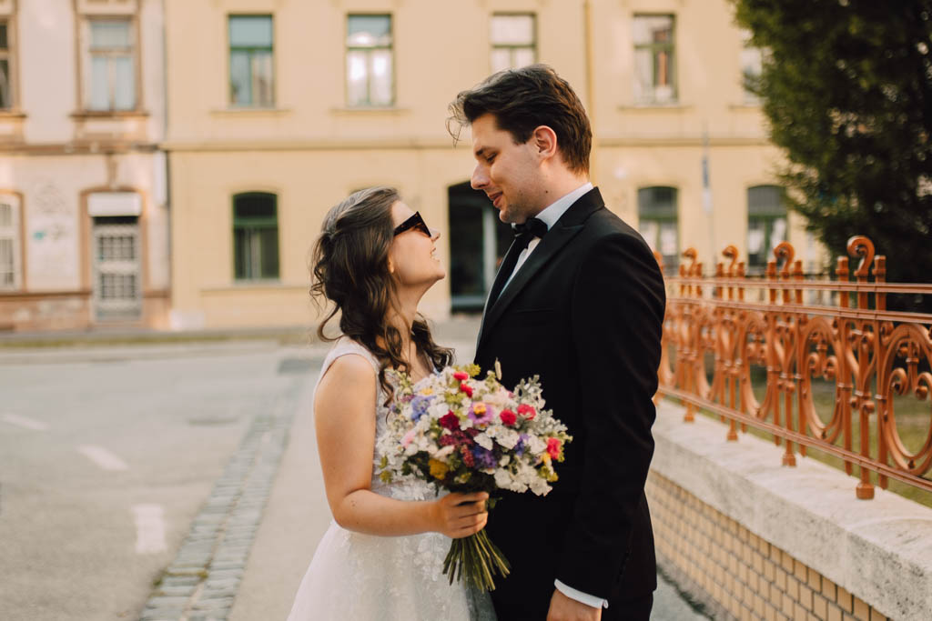 Také pekné - svadobný fotograf - Košice, Prešov Nevesta s farebnou kyticou a ženích v čiernom obleku stoja oproti sebe a usmievajú sa na mestskej ulici s historickými budovami a ozdobným železným plotom v pozadí.