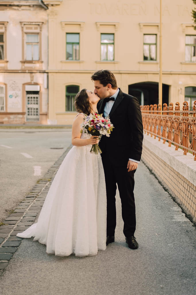 Také pekné - svadobný fotograf - Košice, Prešov Nevesta v bielych šatách bozkáva na líce ženícha v čiernom obleku s motýlikom, ktorý drží farebnú kyticu, ako spolu stoja na tichej ulici s historickými budovami v pozadí.