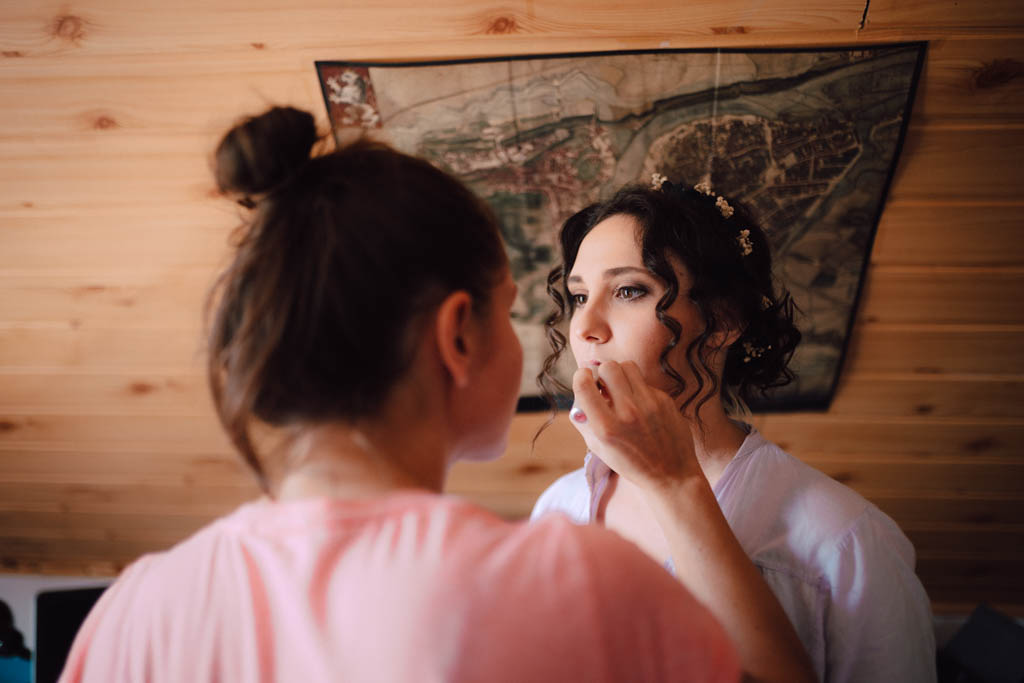 Také pekné - svadobný fotograf - Košice, Prešov Ženu s natočenými tmavými vlasmi a kvetinovými sponkami líči iná žena v ružovej košeli v miestnosti s dreveným obložením a historickou mapou na stene.