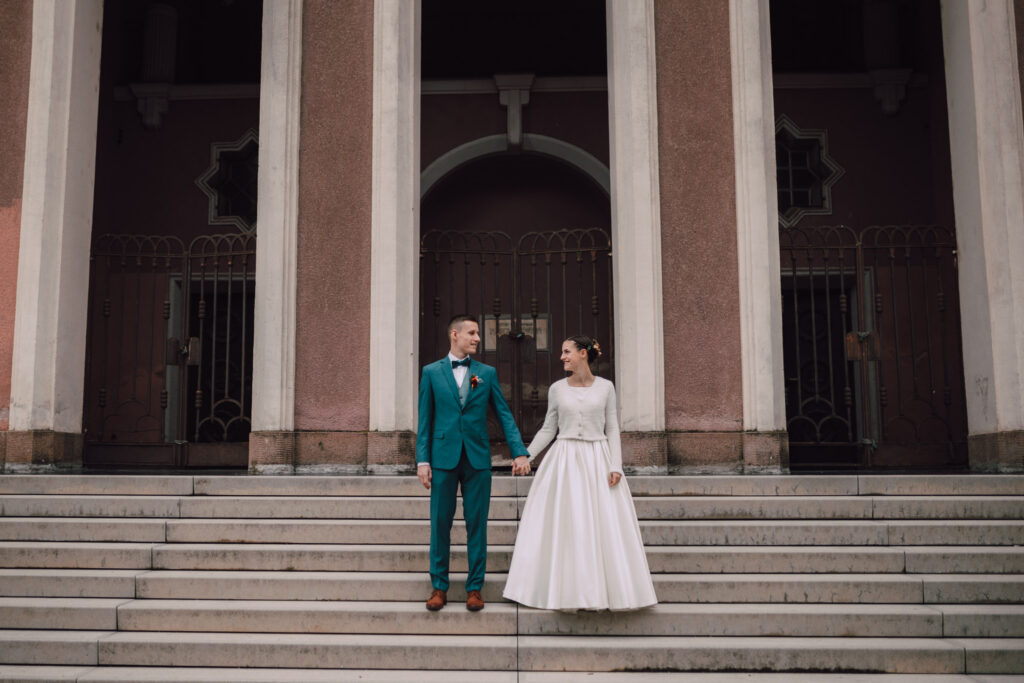 Také pekné - svadobný fotograf - Košice, Prešov Nevesta a ženích sa držia za ruky a stoja na kamenných schodoch pred budovou s vysokými stĺpmi a ružovými stenami. Ženích má oblečený oblek v sýtozelenej farbe a nevesta má oblečené biele šaty s dlhými rukávmi.