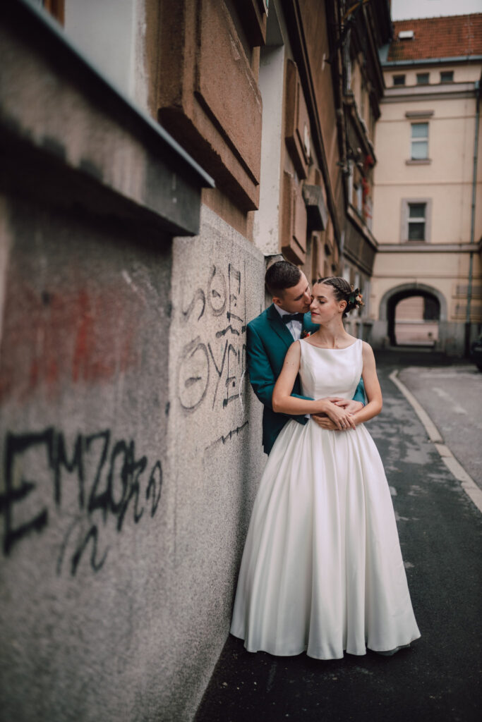 Také pekné - svadobný fotograf - Košice, Prešov Mladomanželia sa objímajú na ulici v meste, ženích má na sebe čajový oblek a nevesta biele šaty. Stoja pri stene s grafitmi a zdieľajú intímnu chvíľu v mestskom prostredí.