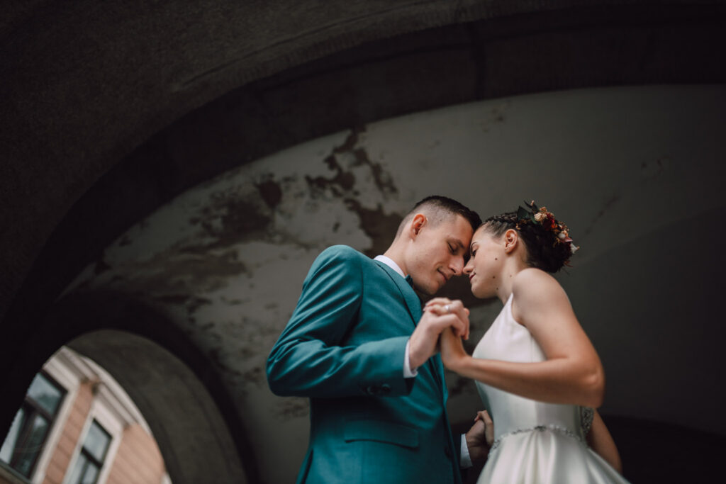 Také pekné - svadobný fotograf - Košice, Prešov Nevesta a ženích stoja blízko seba pod oblúkom, držia sa za ruky a dotýkajú sa čelom. Nevesta má na sebe biele šaty a kvetinovú čelenku, ženích má na sebe oblek v talianskej farbe. Na pozadí je zvetraná architektúra.