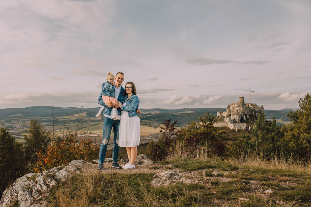 Také pekné - svadobný fotograf - Košice, Prešov Trojčlenná rodina stojí na skalnatom kopci a usmieva sa do kamery. Muž drží malé dieťa a žena stojí vedľa nich. V pozadí je starý hrad a zvlnené kopce pod zamračenou oblohou.