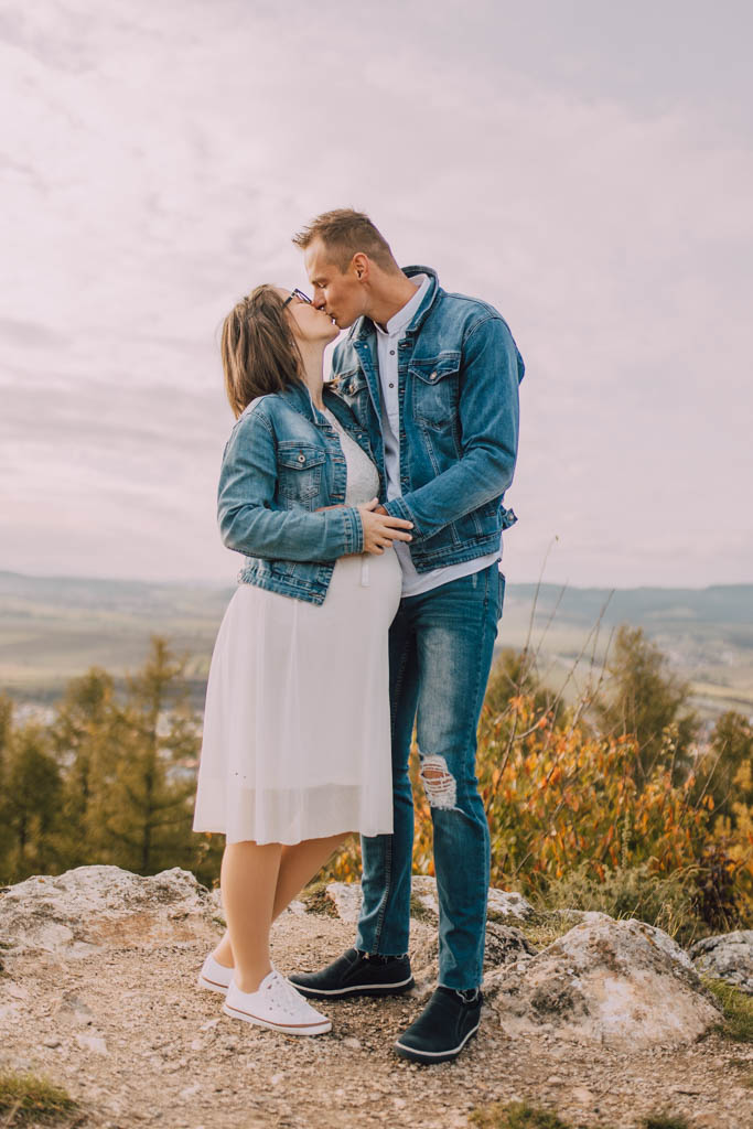 Také pekné - svadobný fotograf - Košice, Prešov Pár stojí vonku na skalnatom kopci a bozkáva sa. Žena má na sebe biele šaty a džínsovú bundu a drží si bruško, zatiaľ čo muž má na sebe džínsy a džínsovú bundu. V pozadí je malebná krajina a zamračená obloha.