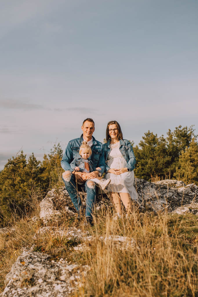 Také pekné - svadobný fotograf - Košice, Prešov Trojčlenná rodina, muž, žena a malé dieťa, sedí na veľkej skale na trávnatom poli so stromami v pozadí. Usmievajú sa a majú na sebe bežné oblečenie. Obloha je jasná.