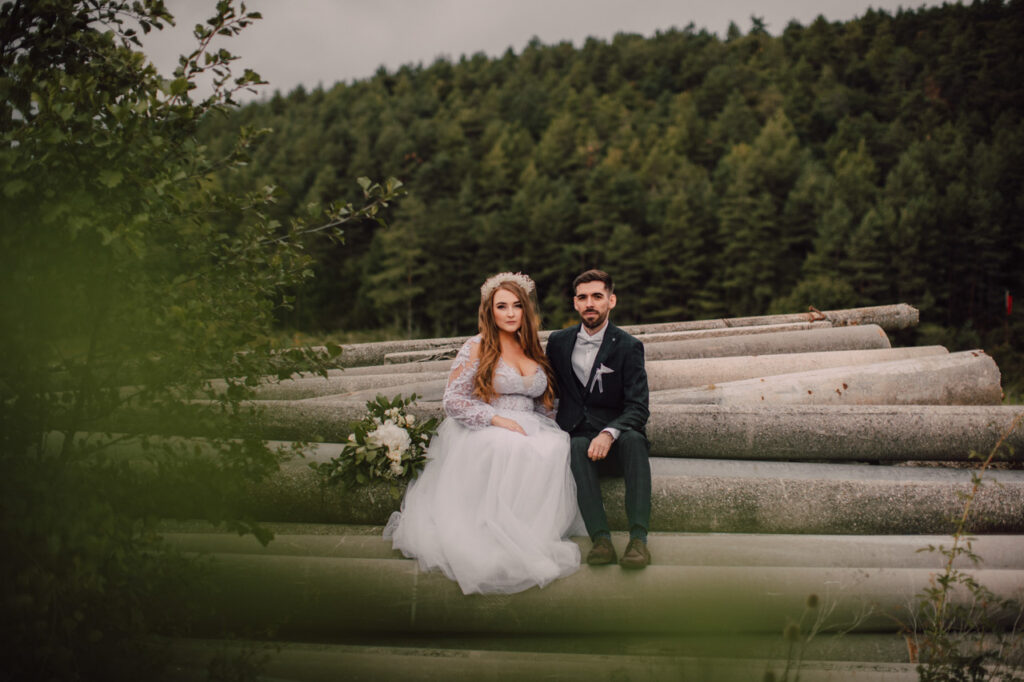 Také pekné - svadobný fotograf - Košice, Prešov Nevesta v bielych šatách a ženích v tmavom obleku sedia spolu na veľkých kládach vonku, obklopení zeleňou a zalesneným úbočím v pozadí.