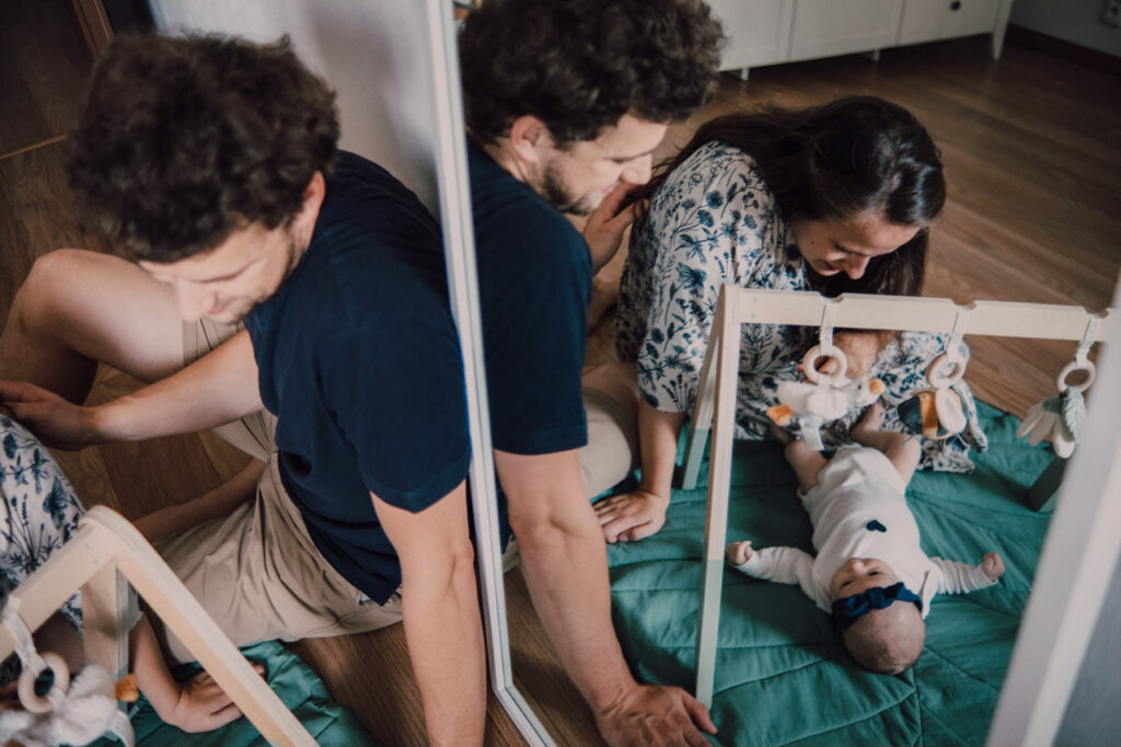 Také pekné - svadobný fotograf - Košice, Prešov Muž a žena sedia na zemi vedľa dieťaťa ležiaceho na podložke a hrajú sa s drevenou detskou posilňovňou. Scéna sa odráža v zrkadle vedľa nich. Nálada je teplá a hravá.
