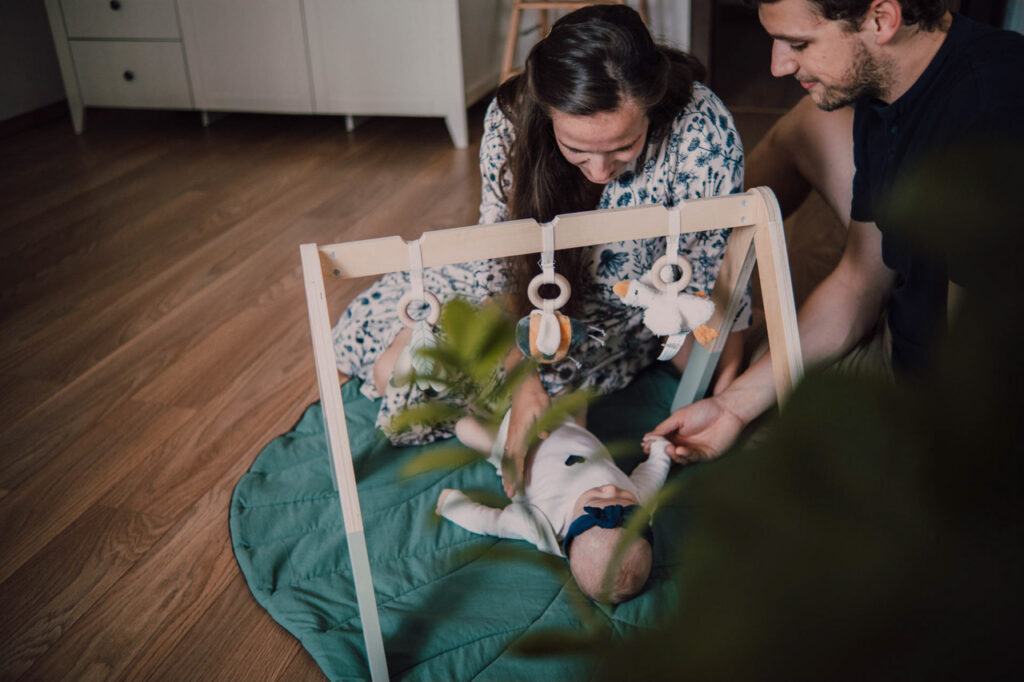 Také pekné - svadobný fotograf - Košice, Prešov Dieťa leží na zelenej podložke pod drevenou hracou plošinou so zavesenými hračkami, zatiaľ čo dvaja dospelí, pravdepodobne rodičia, sedia neďaleko na zemi, komunikujú a usmievajú sa na dieťa.