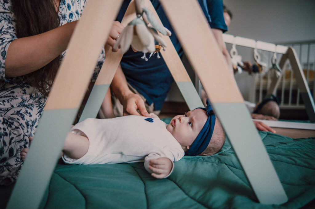 Také pekné - svadobný fotograf - Košice, Prešov Dieťa leží na zelenej podložke pod drevenou hracou plošinou a pozerá sa na zavesené hračky. Dospelý pomáha upravovať hračky nad dieťaťom v útulnom interiéri.