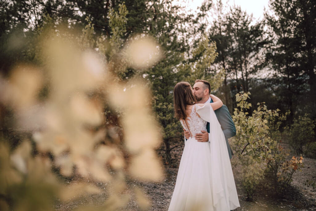 Také pekné - svadobný fotograf - Košice, Prešov Nevesta a ženích sa objímajú a bozkávajú pod holým nebom v lesnom prostredí, obklopení stromami a jemným, rozmazaným lístím v popredí. Scéna je teplá a romantická.