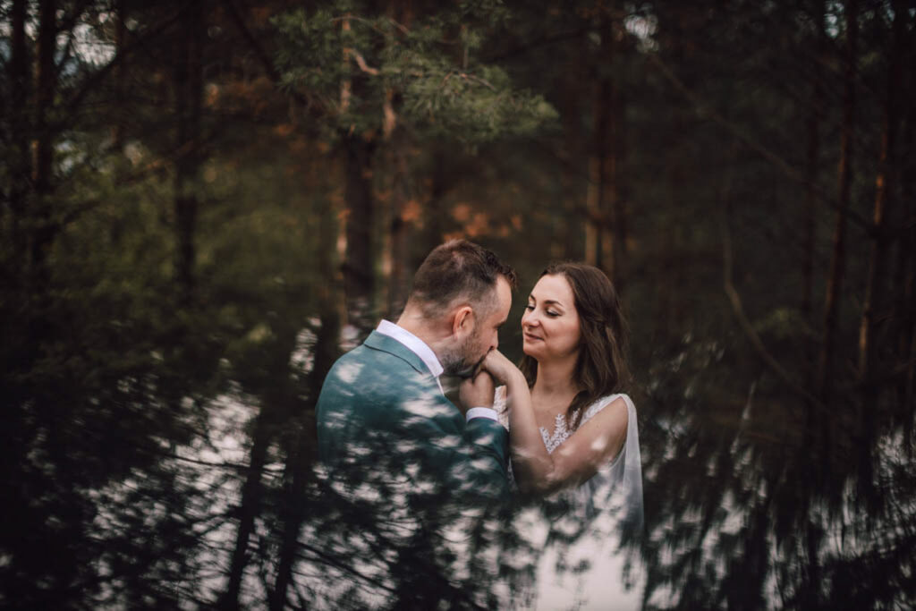 Také pekné - svadobný fotograf - Košice, Prešov Muž v obleku jemne drží a bozkáva ruku ženy, ktorá sa usmieva so zatvorenými očami. Stoja blízko seba v lese, obklopení vysokými stromami a mäkkým prirodzeným svetlom.