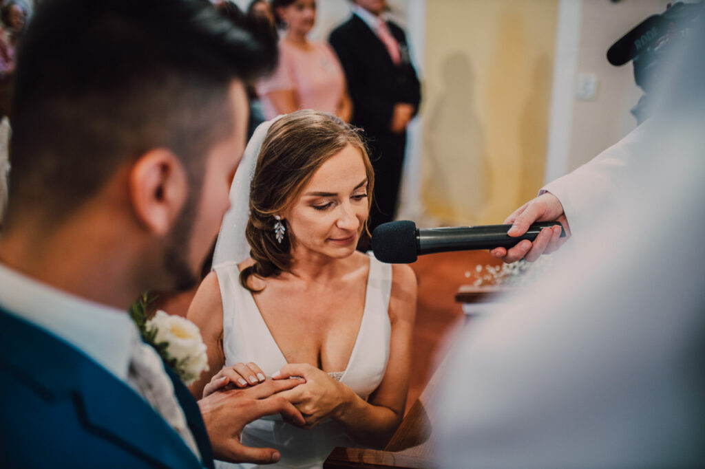 Také pekné - svadobný fotograf - Košice, Prešov Nevesta drží ženícha za ruku a hovorí do mikrofónu počas svadobného obradu. Ženích je čiastočne viditeľný a ostatní ľudia v pozadí sú rozmazaní.
