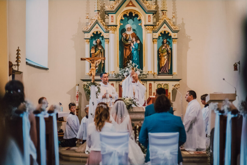 Také pekné - svadobný fotograf - Košice, Prešov Svadobný obrad v kostole, pri ktorom kňaz slúži pri oltári, nevesta a ženích kľačia pred oltárom a hostia a družbovia sú oblečení v slávnostných šatách. Zdobený oltár je vyzdobený sochami a kvetmi.
