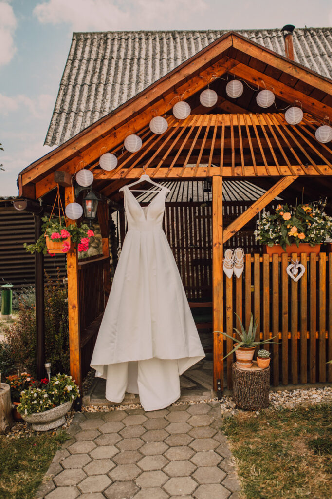 Také pekné - svadobný fotograf - Košice, Prešov Biele svadobné šaty visia na drevenej konštrukcii ozdobenej papierovými lampiónmi a kvetmi, v popredí je rustikálna záhrada a kamenná cestička.